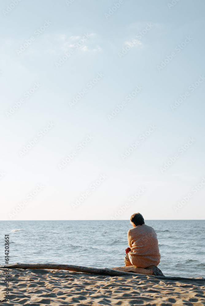 Lonely pensive adult woman sitting alone on a log on the seashore looking at the sea, view from the back