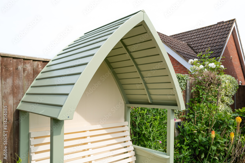 Fototapeta premium New installed wooden contemporary arbour showing its ornate roof structure and two seat arrangement. Seen against a neighbouring fence.