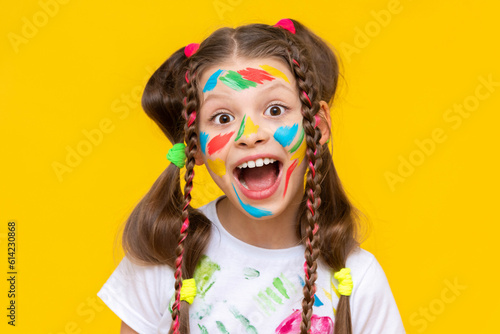 Funny little girl with a painted face with colorful paints. Happy childhood. Isolated yellow background.