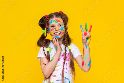 A young girl stained in multicolored paint shows three fingers and smiles broadly. Children's creativity. Yellow isolated background.