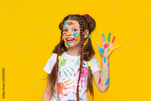 A young girl stained in multicolored paint shows five fingers and smiles broadly. Children s creativity. Yellow isolated background.