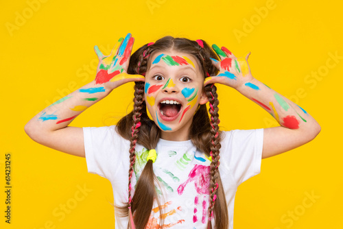A young girl with brightly painted hands and multi colored pigtails shows her palms, smiling broadly. Yellow isolated background.