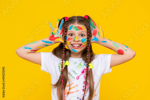 A young girl with brightly painted hands and multi colored pigtails shows her palms, smiling broadly. Yellow isolated background.