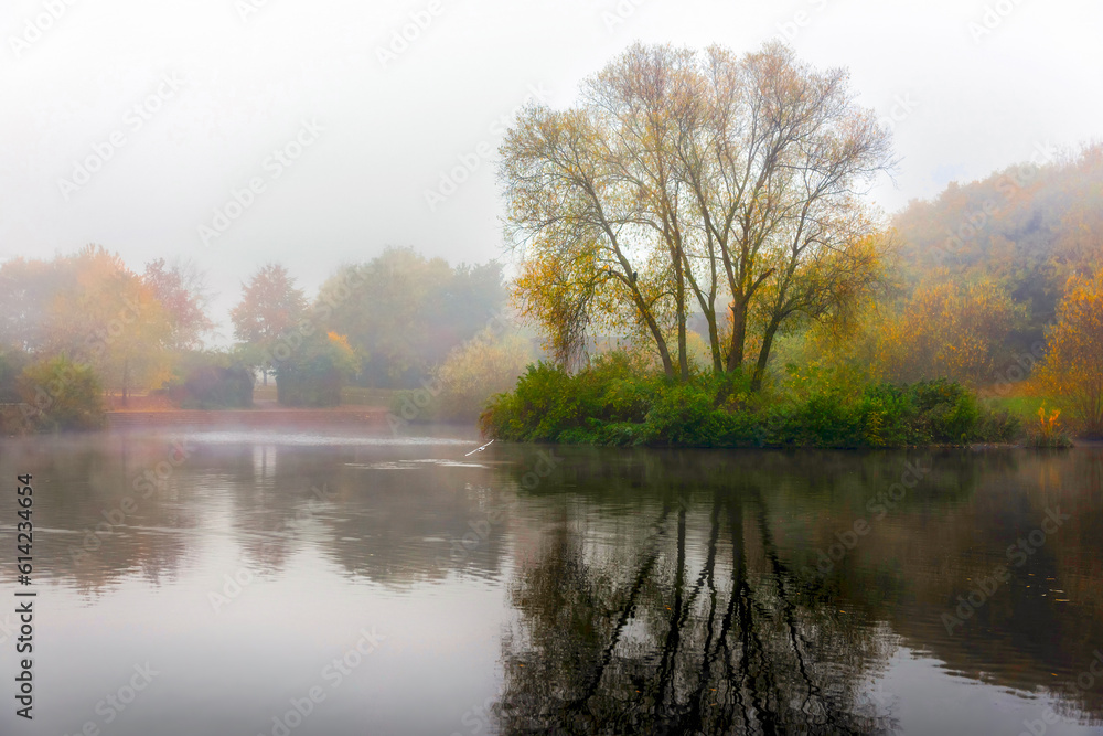 View of foggy lake, island and autumn color trees with reflection on water and copy space in clear sky.