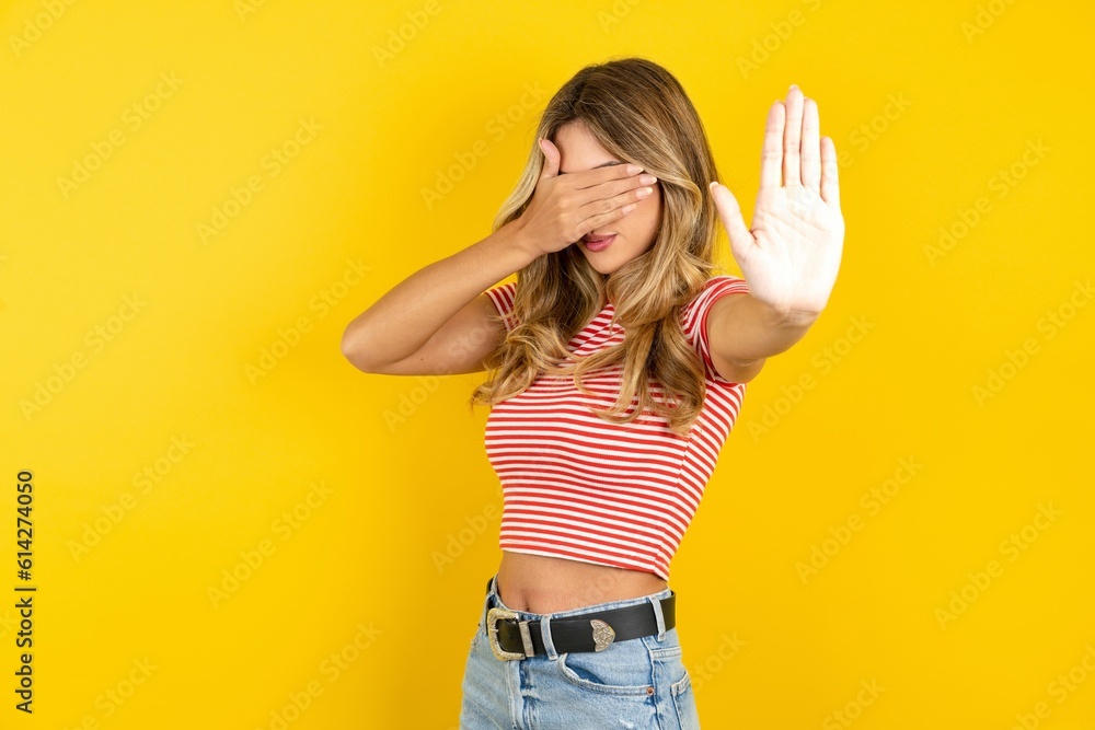 Young beautiful woman wearing striped t-shirt over yellow background covers eyes with palm and doing stop gesture, tries to hide from everybody. Young beautiful woman wearing striped t-shirt over yellow background covers eyes with palm and doing stop gesture, tries to hide from everybody.