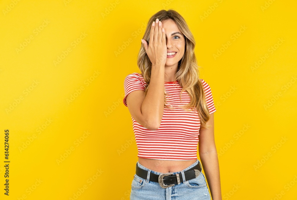 Young beautiful woman wearing striped t-shirt over yellow background covering one eye with her hand, confident smile on face and surprise emotion. Young beautiful woman wearing striped t-shirt over yellow background covering one eye with her hand, confident smile on face and surprise emotion.