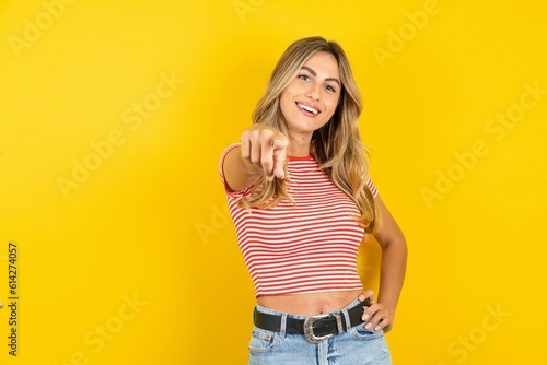 Young beautiful woman wearing striped t-shirt over yellow background pointing at camera with a satisfied, confident, friendly smile, choosing you