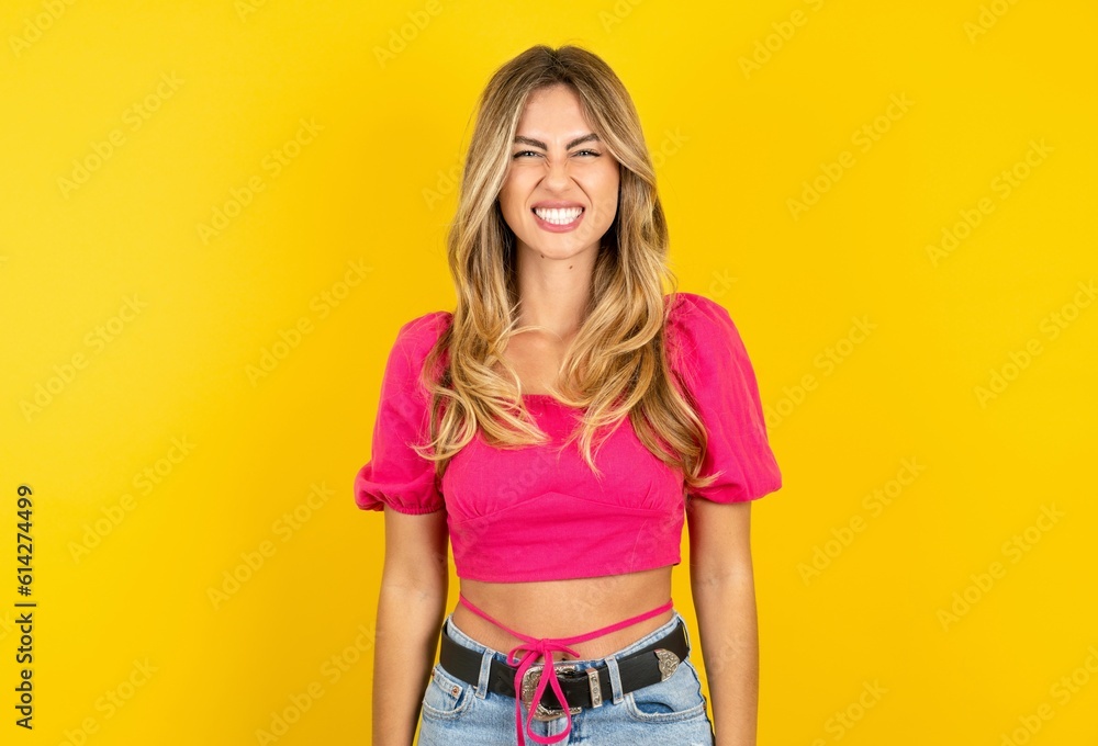 Young beautiful blonde woman wearing pink crop top over yellow studio background keeps teeth clenched, frowns face in dissatisfaction, irritated because of much duties. Young beautiful blonde woman wearing pink crop top over yellow studio background keeps teeth clenched, frowns face in dissatisfaction, irritated because of much duties.