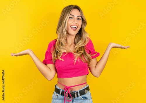 Cheerful cheery optimistic Young beautiful blonde woman wearing pink crop top over yellow studio background holding two palms copy space