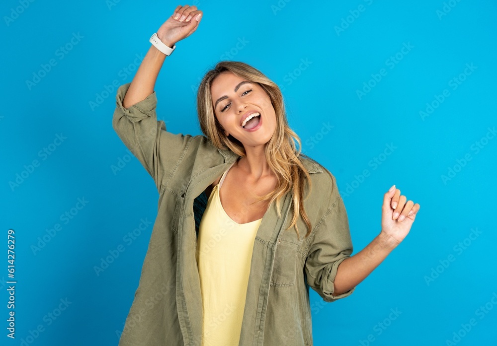 Fototapeta premium Photo of upbeat young beautiful blonde woman standing over blue studio background has fun and dances carefree wear being in perfect mood makes movements. Spends free time on disco party