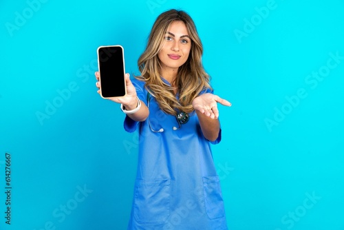 Young beautiful doctor woman standing over blue studio background with a mobile. presenting smartphone. Advertisement concept.