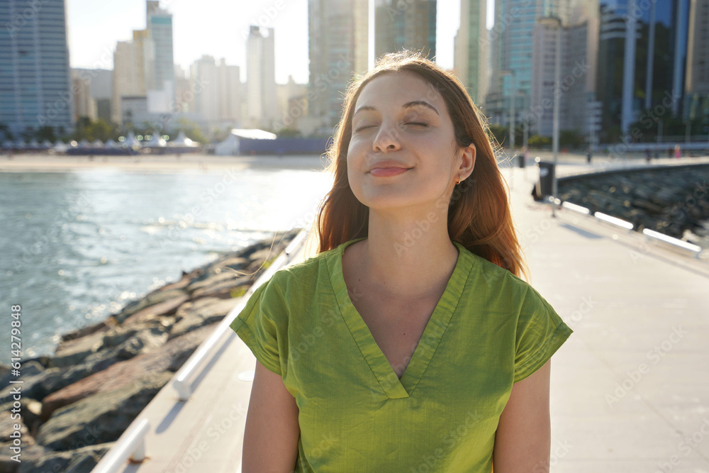 Portrait of beautiful young Brazilian woman with closed eyes enjoying sun on city promenade at sunset