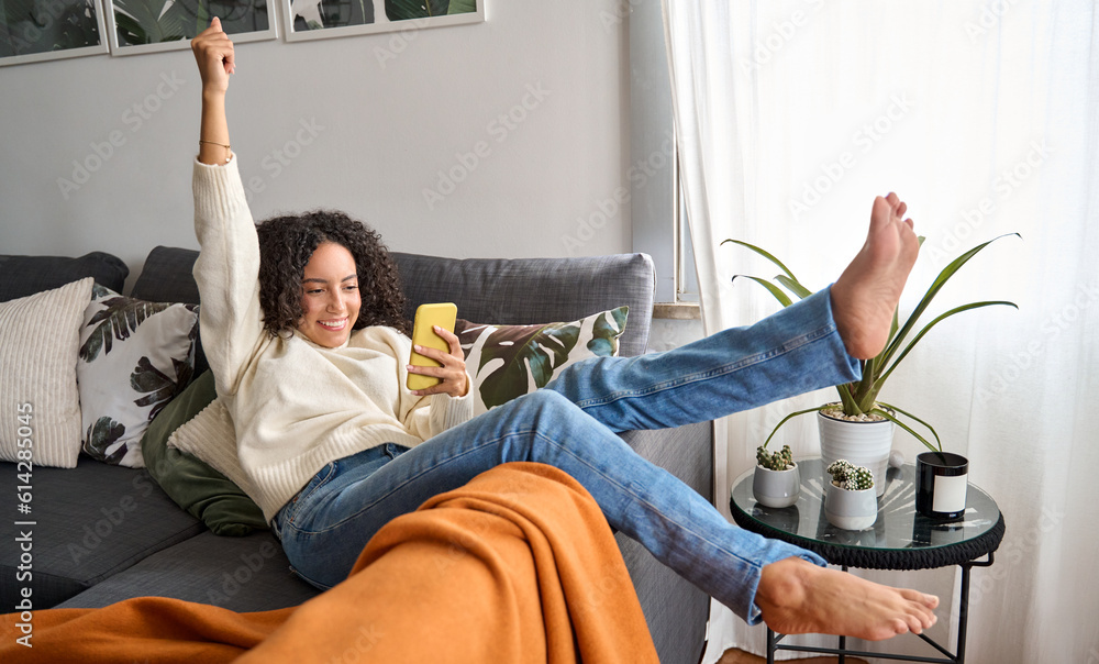 Happy excited young latin woman relaxing on couch using phone winning money in online app game. Young lucky woman feeling winner looking at cellphone, receiving great news or discount offer. - obrazy, fototapety, plakaty 