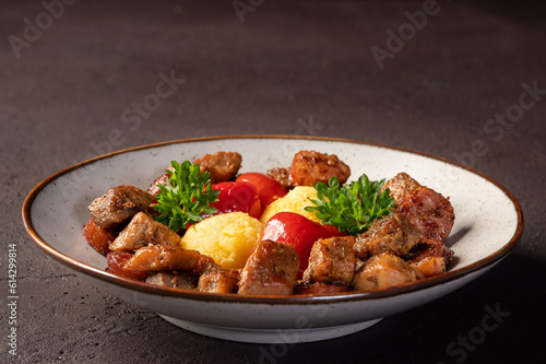 Romanian festive dish pomana porcului consisting of pieces of fried pork and sausages with pickled red peppers and polenta close-up on a plate on dark background. 
