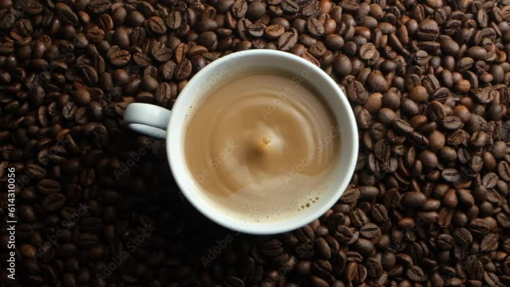 Top view, flat lay of a cup of freshly brewed delicious coffee is placed on the background of brown roasted coffee beans, a drop of coffee falls into the glass and the water splashes up slowly. Top view, flat lay of a cup of freshly brewed delicious coffee is placed on the background of brown roasted coffee beans, a drop of coffee falls into the glass and the water splashes up slowly.