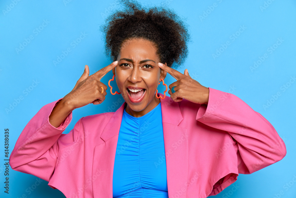 Naklejka premium Portrait of beautiful black girl against blue background, having good time in studio, pressing fingers to her head and smiles, delight concept, copy space