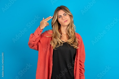 Unhappy young beautiful blonde woman wearing overshirt makes suicide gesture and imitates gun with hand, curves lips, keeps two fingers on temple