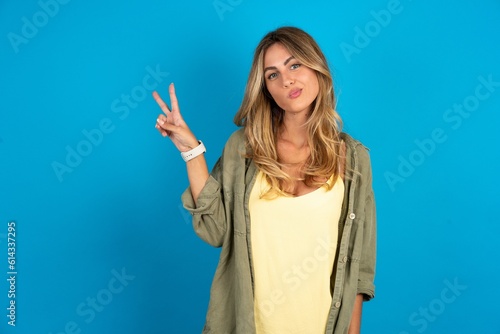 young beautiful blonde woman wearing overshirt makes peace gesture keeps lips folded shows v sign. Body language concept