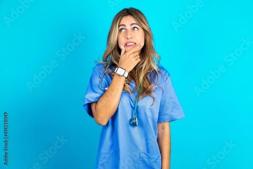 Young caucasian blonde doctor woman wearing blue uniform Thinking worried about a question, concerned and nervous with hand on chin.