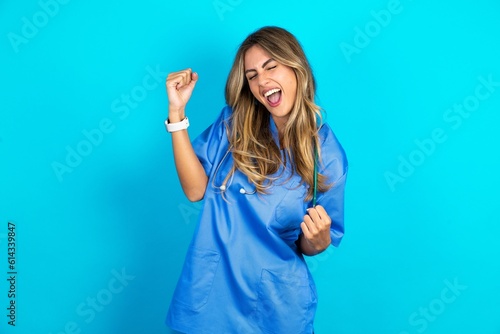 Young caucasian blonde doctor woman wearing blue uniform very happy and excited doing winner gesture with arms raised, smiling and screaming for success. Celebration concept.