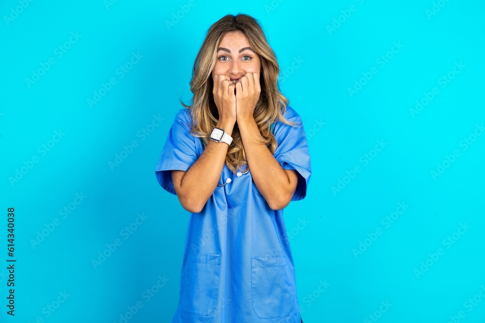 Fearful Young caucasian blonde doctor woman wearing blue uniform keeps hands near mouth, feels frightened and scared, has a phobia, Shock and frighted concept. Fearful Young caucasian blonde doctor woman wearing blue uniform keeps hands near mouth, feels frightened and scared, has a phobia, Shock and frighted concept.