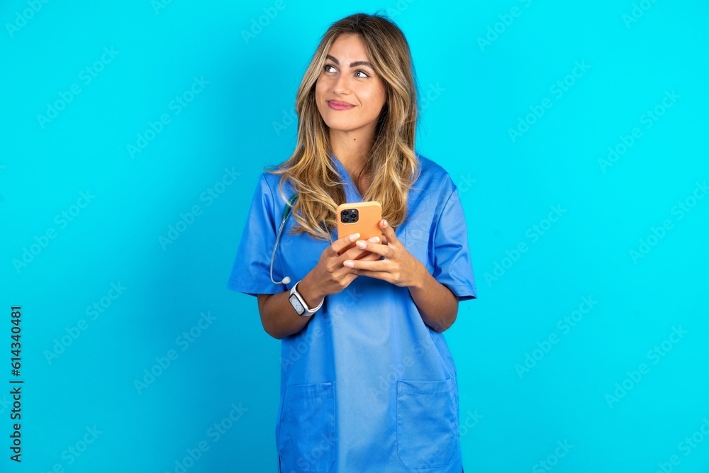 Young caucasian blonde doctor woman wearing blue uniform holding in hands mobile phone and looking aside with dreamy look.