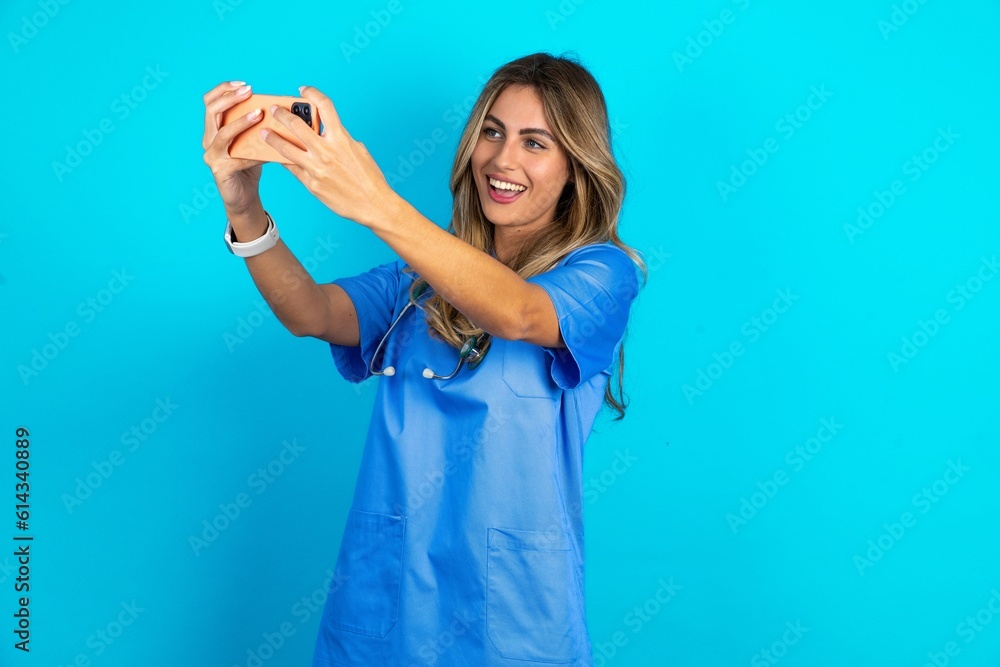 Fototapeta premium Young caucasian blonde doctor woman wearing blue uniform taking a selfie to post it on social media or having a video call with friends.