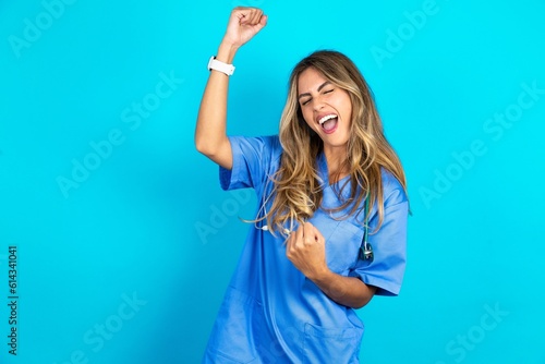Profile photo of excited Young caucasian blonde doctor woman wearing blue uniform good mood raise fists screaming rejoicing overjoyed basketball sports fan supporter