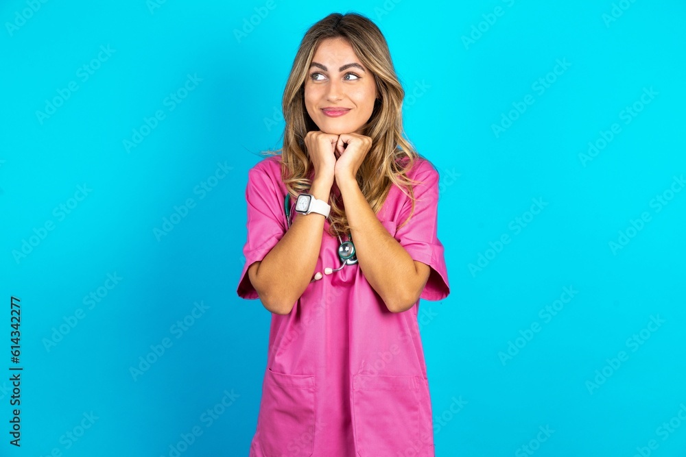 Happy Young caucasian blonde woman wearing pink uniform anticipates something awesome happen, looks happily aside, keeps hands together near face, has glad expression. Happy Young caucasian blonde woman wearing pink uniform anticipates something awesome happen, looks happily aside, keeps hands together near face, has glad expression.