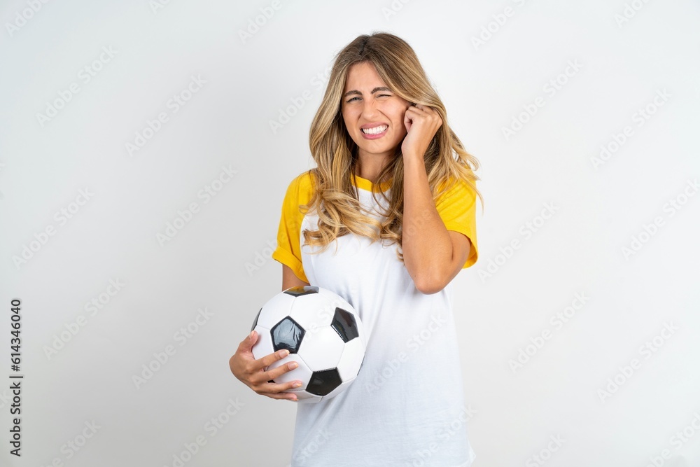 Happy Young beautiful woman wearing football T-shirt over white background ignores loud music and plugs ears with fingers asks to turn off sound Happy Young beautiful woman wearing football T-shirt over white background ignores loud music and plugs ears with fingers asks to turn off sound