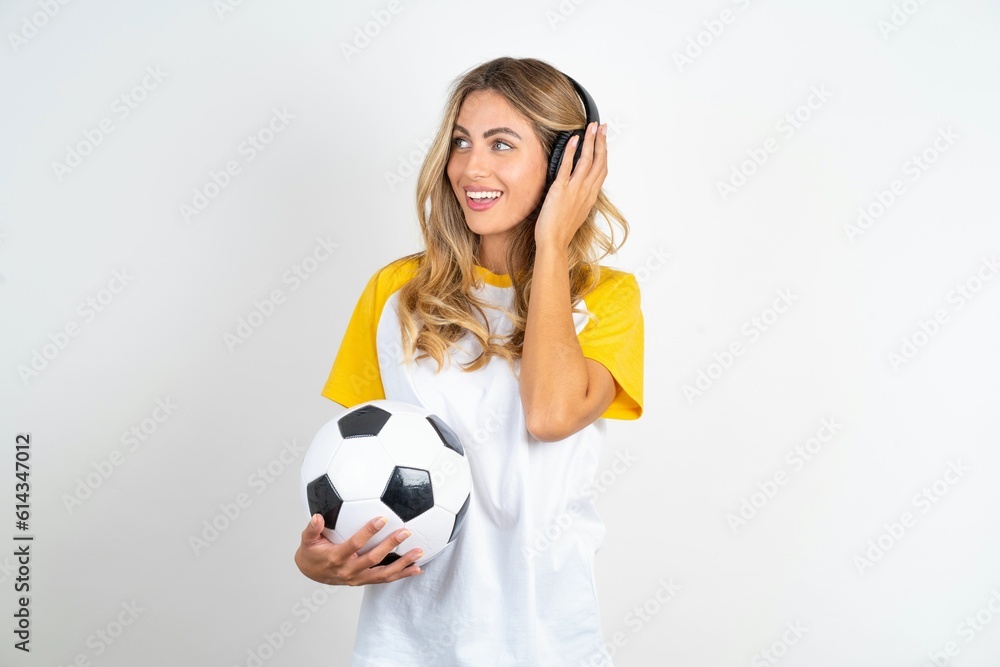 Young beautiful woman wearing football T-shirt over white background wears stereo headphones listens music concentrated aside. People hobby lifestyle concept Young beautiful woman wearing football T-shirt over white background wears stereo headphones listens music concentrated aside. People hobby lifestyle concept