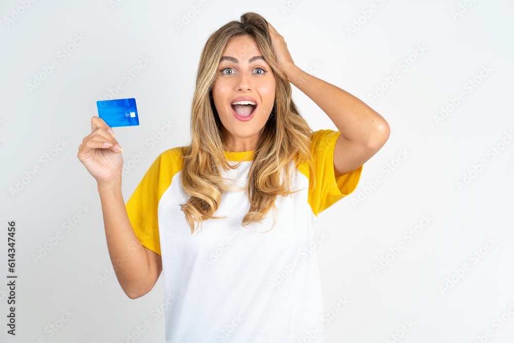 Impressed funny Young beautiful woman wearing football T-shirt over white background hand on head holding bank card Impressed funny Young beautiful woman wearing football T-shirt over white background hand on head holding bank card