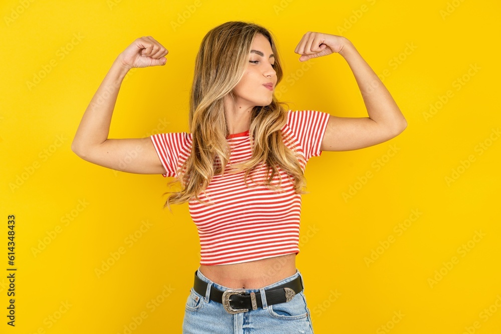 Young beautiful blonde woman wearing striped t-shirt over yellow studio background showing arms muscles smiling proud. Fitness concept. Young beautiful blonde woman wearing striped t-shirt over yellow studio background showing arms muscles smiling proud. Fitness concept.