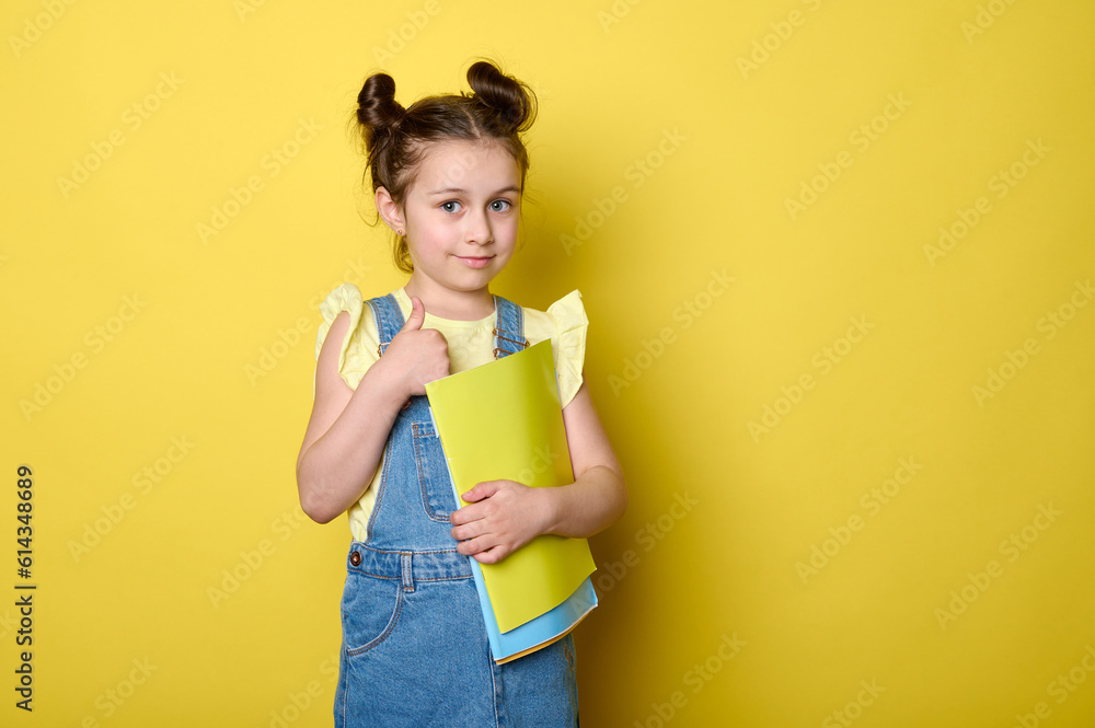 Happy smart schoolgirl, first grader gesturing with thumb up, smiling looking at camera, feeling happy to start a new semester in new primary school, isolated yellow studio background with copy space Happy smart schoolgirl, first grader gesturing with thumb up, smiling looking at camera, feeling happy to start a new semester in new primary school, isolated yellow studio background with copy space