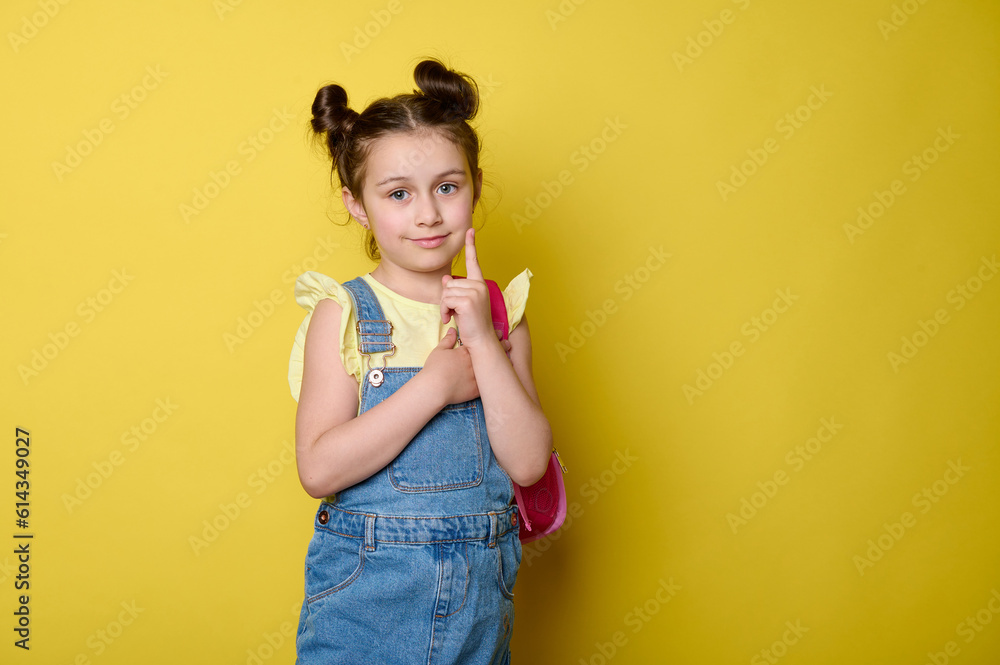 Stylish cute little girl 6 years old, primary school student in casual denim, carrying a pink rucksack, showing index finger, smiling looking at camera, isolated on yellow background with copy space Stylish cute little girl 6 years old, primary school student in casual denim, carrying a pink rucksack, showing index finger, smiling looking at camera, isolated on yellow background with copy space