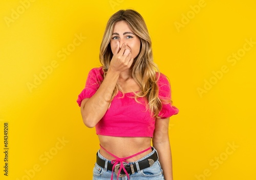 young blonde woman wearing pink crop top over yellow studio background smelling something stinky and disgusting, intolerable smell, holding breath with fingers on nose. Bad smell