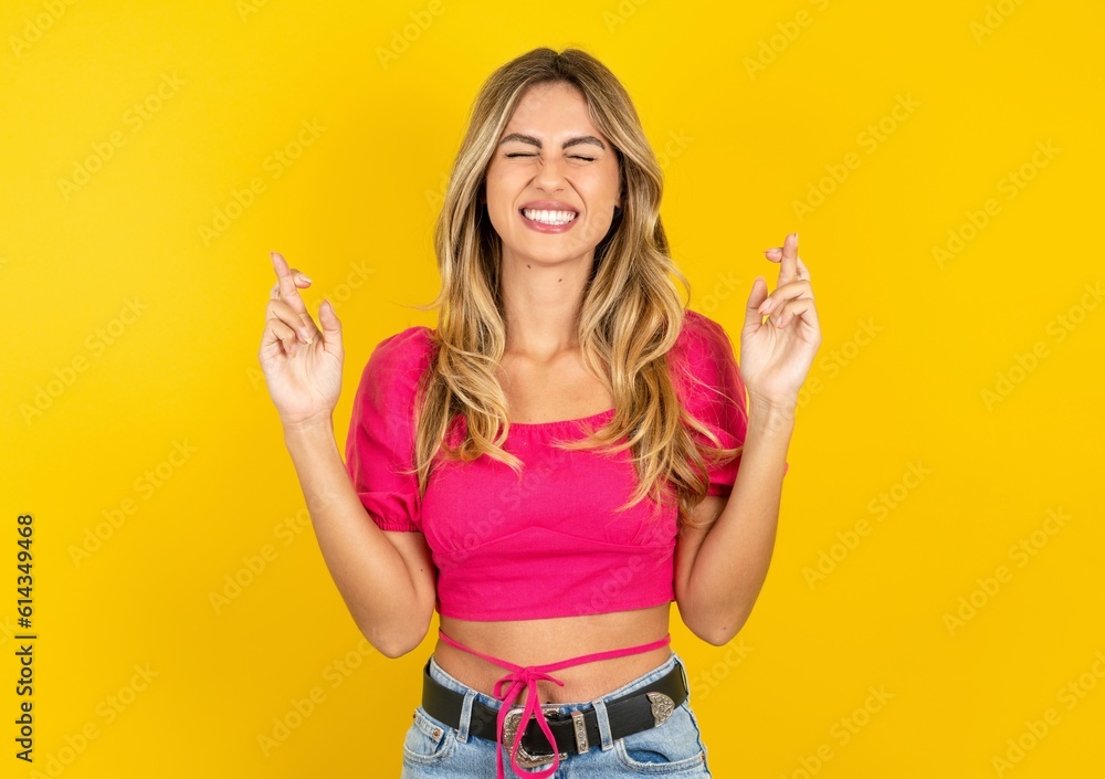 young blonde woman wearing pink crop top over yellow studio background gesturing finger crossed smiling with hope and eyes closed. Luck and superstitious concept. young blonde woman wearing pink crop top over yellow studio background gesturing finger crossed smiling with hope and eyes closed. Luck and superstitious concept.