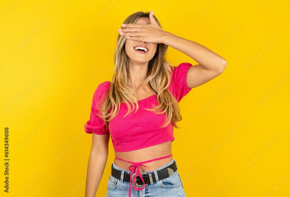 young blonde woman wearing pink crop top over yellow studio background smiling and laughing with hand on face covering eyes for surprise. Blind concept. young blonde woman wearing pink crop top over yellow studio background smiling and laughing with hand on face covering eyes for surprise. Blind concept.