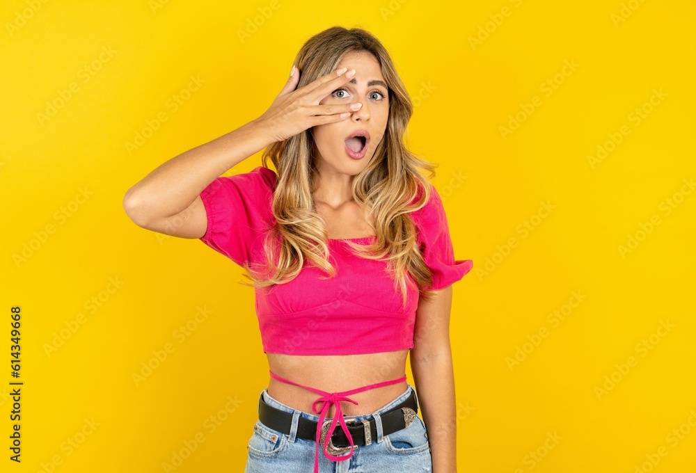 young blonde woman wearing pink crop top over yellow studio background peeking in shock covering face and eyes with hand, looking through fingers with embarrassed expression. young blonde woman wearing pink crop top over yellow studio background peeking in shock covering face and eyes with hand, looking through fingers with embarrassed expression.