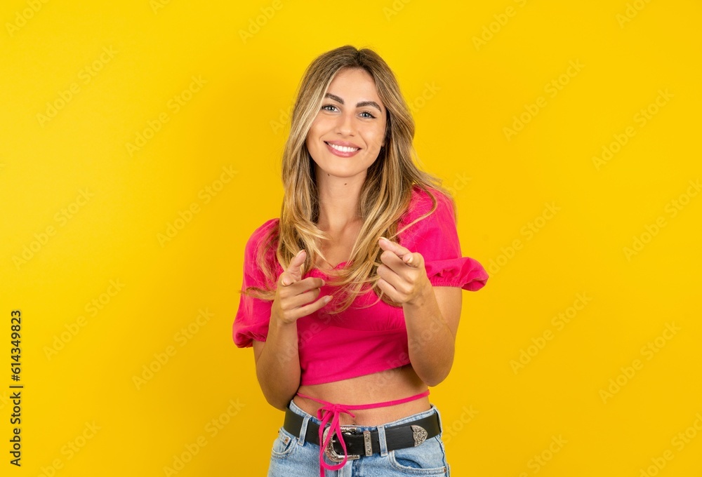 young blonde woman wearing pink crop top over yellow studio background pointing fingers to camera with happy and funny face. Good energy and vibes. young blonde woman wearing pink crop top over yellow studio background pointing fingers to camera with happy and funny face. Good energy and vibes.