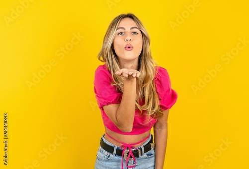 young blonde woman wearing pink crop top over yellow studio background looking at the camera blowing a kiss with hand on air being lovely and sexy. Love expression.