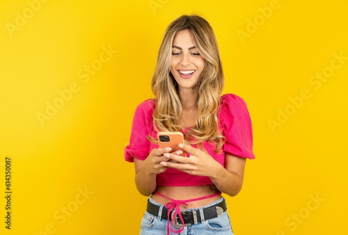 Fotografia young blonde woman wearing pink crop top over yellow studio background using mobile phone chatting free time