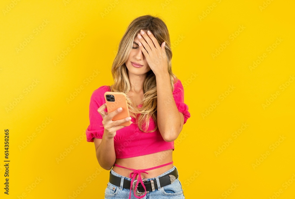 young blonde woman wearing pink crop top over yellow studio background looking at smart phone feeling sad holding hand on face. young blonde woman wearing pink crop top over yellow studio background looking at smart phone feeling sad holding hand on face.