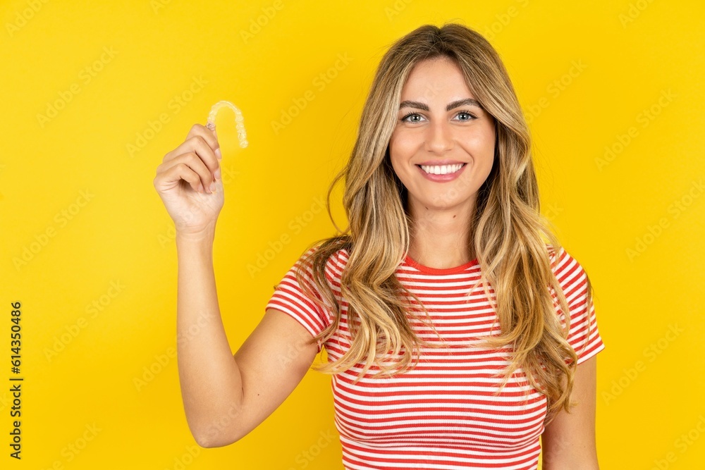 Young beautiful blonde woman wearing striped t-shirt over yellow studio background holding an invisible braces aligner, recommending this new treatment. Dental healthcare concept. Young beautiful blonde woman wearing striped t-shirt over yellow studio background holding an invisible braces aligner, recommending this new treatment. Dental healthcare concept.