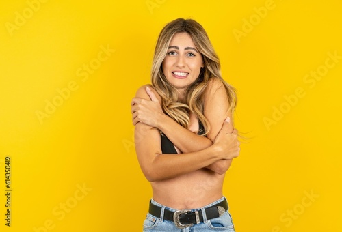 Desperate young beautiful blonde woman wearing bikini over yellow studio background trembles and feels cold, hugs oneself to warm up or feels scared notices something terrifying.
