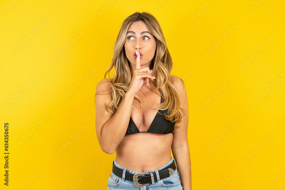 young beautiful blonde woman wearing bikini over yellow studio background silence gesture keeps index finger to lips makes hush sign. Asks not to share secret young beautiful blonde woman wearing bikini over yellow studio background silence gesture keeps index finger to lips makes hush sign. Asks not to share secret