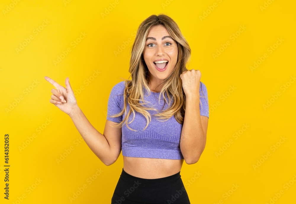 Young beautiful blonde woman wearing sportswear over yellow studio background points at empty space holding fist up, winner gesture. Young beautiful blonde woman wearing sportswear over yellow studio background points at empty space holding fist up, winner gesture.