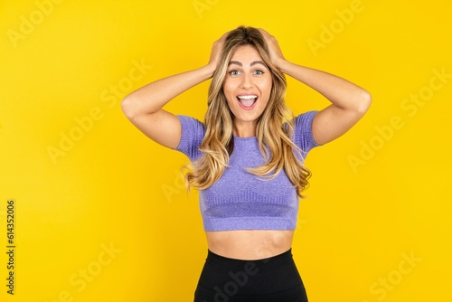 Cheerful overjoyed Young beautiful blonde woman wearing sportswear over yellow studio background reacts rising hands over head after receiving great news.