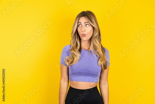 Shocked Young beautiful blonde woman wearing sportswear over yellow studio background look empty space with open mouth screaming: Oh My God! I can't believe this.