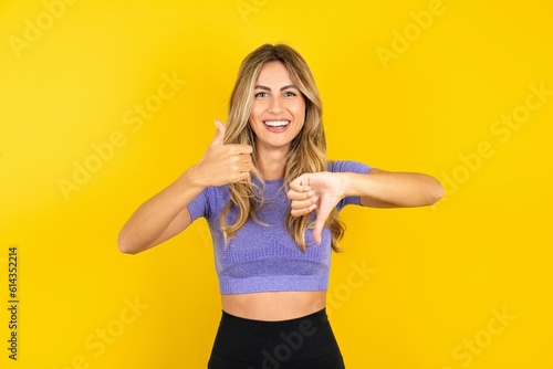 Young beautiful blonde woman wearing sportswear over yellow studio background showing thumb up down sign